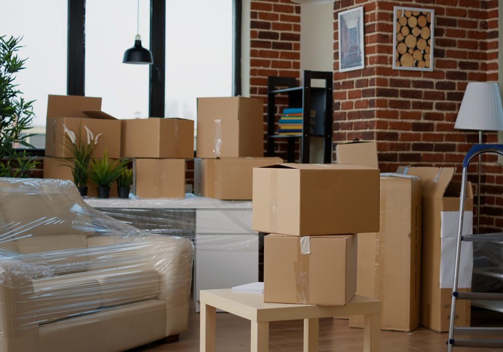 Nobody in empty living room with cardboard packaging at new home, furniture things in stack of carton boxes. No people in household property with package cargo to move in, real estate.